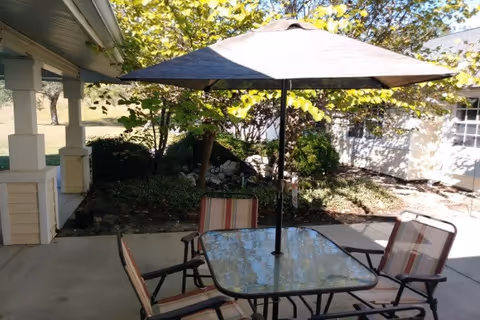 Outdoor patio area with a glass-top table and four striped cushioned chairs under a large umbrella. The patio is adjacent to a building with light-colored siding and surrounded by trees and shrubbery.