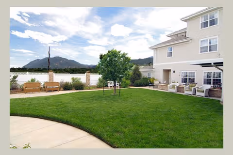 A well-maintained outdoor garden area at Bear Creek Assisted & Senior Living with green grass, a small tree in the center, benches along a white fence, and a multi-story building with patio seating on the right. Mountains and a partly cloudy sky are visible in the background.