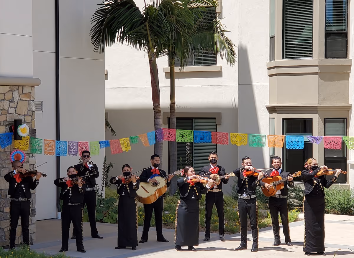 A mariachi band dressed in traditional black outfits with red ties performing outdoors in front of a building. Colorful papel picado banners are strung above them, and there are palm trees and plants in the background.