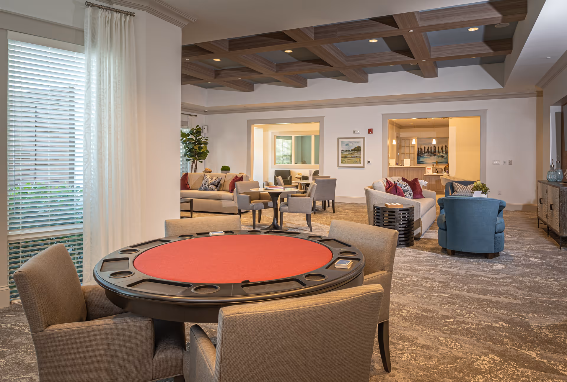 A spacious and well-lit common area in a senior living facility featuring a round poker table with beige chairs in the foreground. In the background, there are multiple seating areas with sofas and armchairs, decorative pillows, a large window with white curtains, a coffered ceiling with recessed lighting, and a reception desk area with pendant lights.