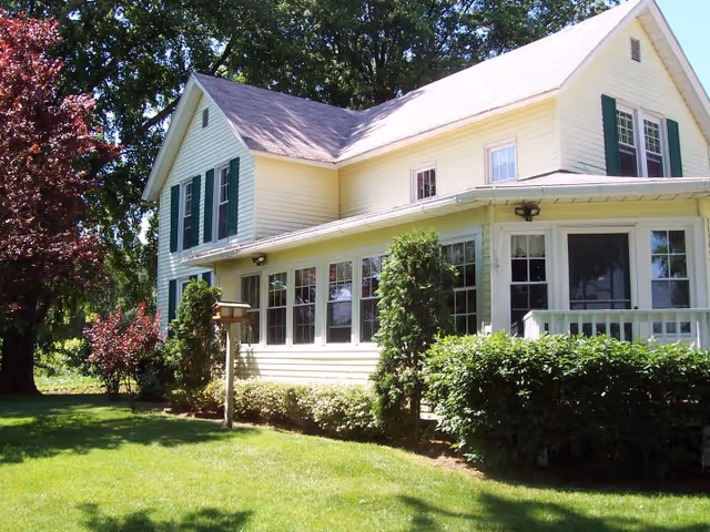 A two-story yellow house with green shutters surrounded by green bushes and trees under a clear blue sky.