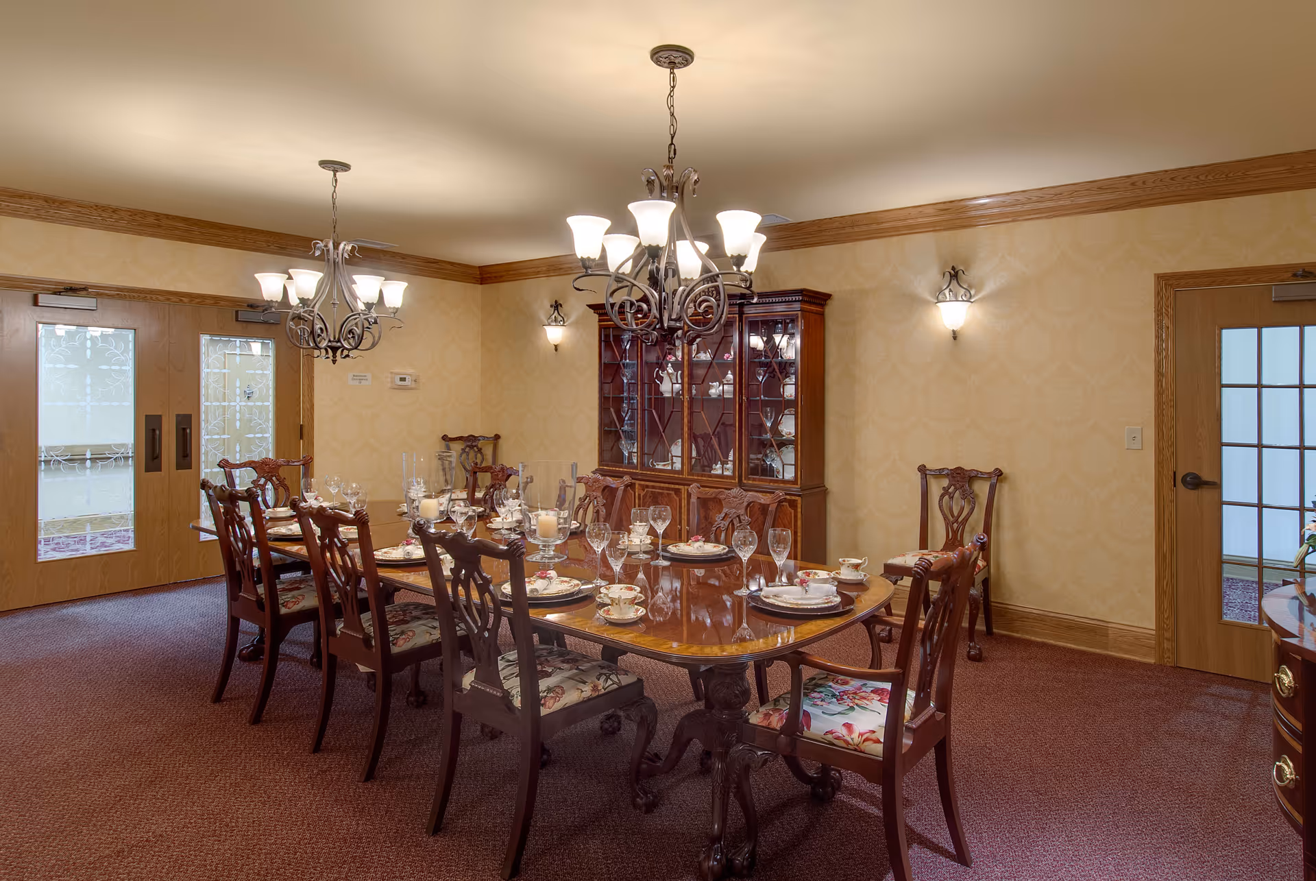 A formal dining room with a long wooden table set for a meal, surrounded by eight wooden chairs with floral cushions. The room features two chandeliers, a wooden china cabinet filled with glassware, beige patterned wallpaper, and two wooden doors with frosted glass panels.