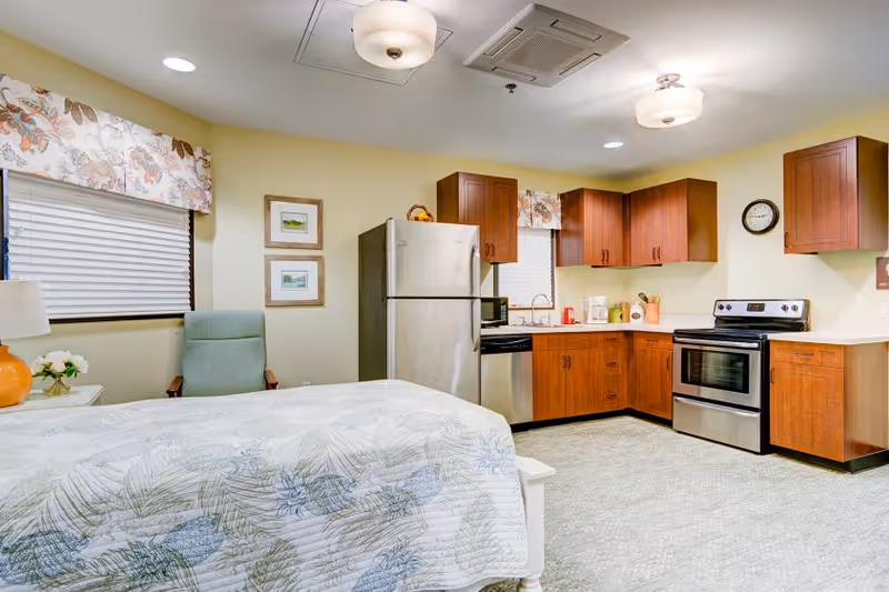 A combined living space featuring a bed with a floral patterned quilt, a green armchair, and a kitchen area with wooden cabinets, stainless steel refrigerator, dishwasher, and stove. The room has light yellow walls, a window with floral valances, two framed pictures, and ceiling lights.