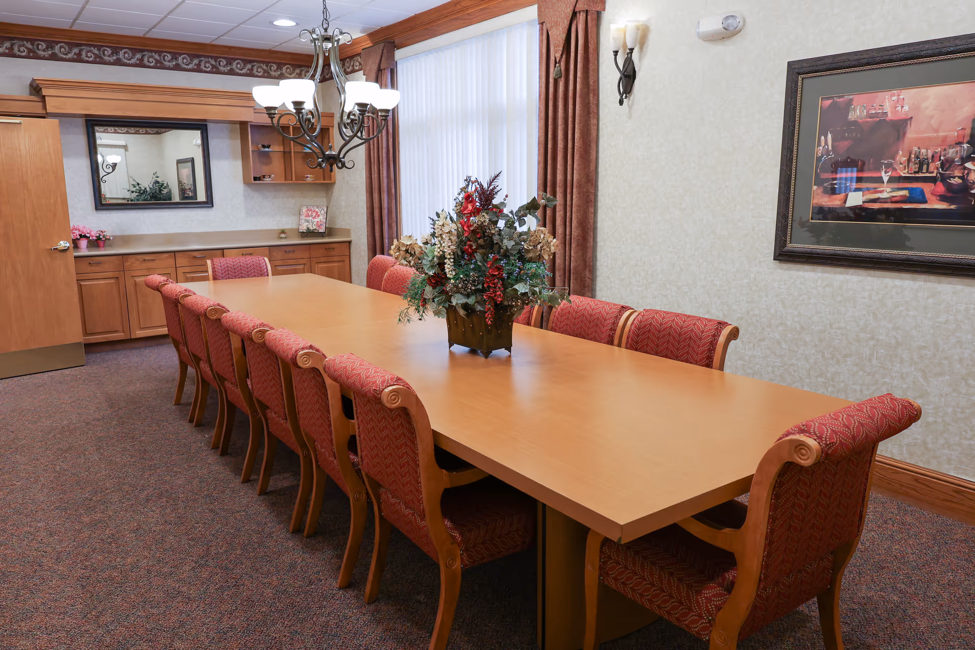A formal dining room with a long wooden table surrounded by twelve red upholstered chairs. A floral centerpiece is placed in the middle of the table. The room has beige walls, a large window with vertical blinds and brown curtains, a chandelier hanging above the table, a framed painting on the wall, and a wooden cabinet with a mirror on the far side.