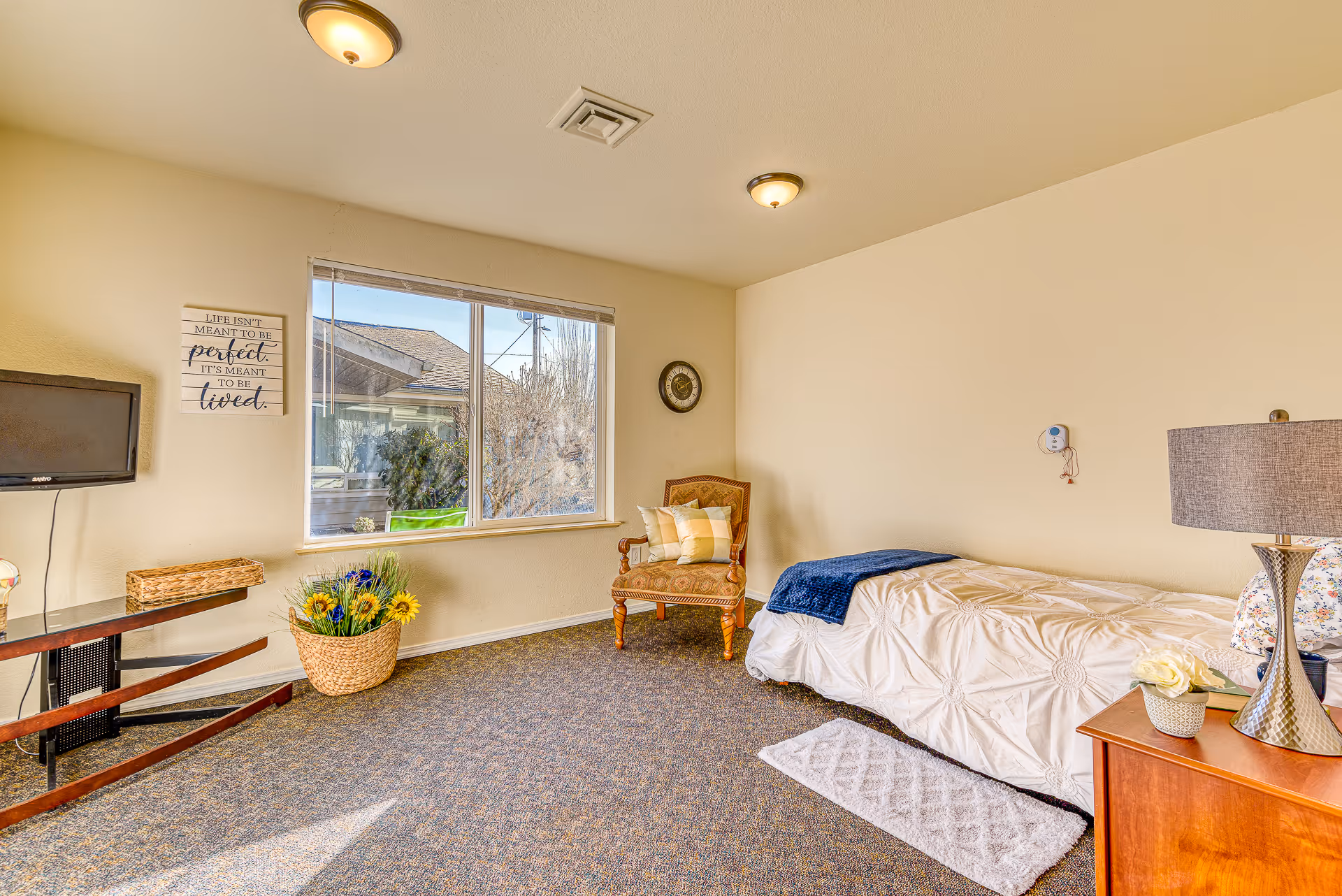 A bright and cozy bedroom in a retirement community featuring a single bed with white bedding and a blue throw blanket, a wooden nightstand with a lamp and a small flower pot, a cushioned armchair with pillows, a wall clock, a flat-screen TV mounted on the wall, a basket with artificial sunflowers, and a large window letting in natural light.