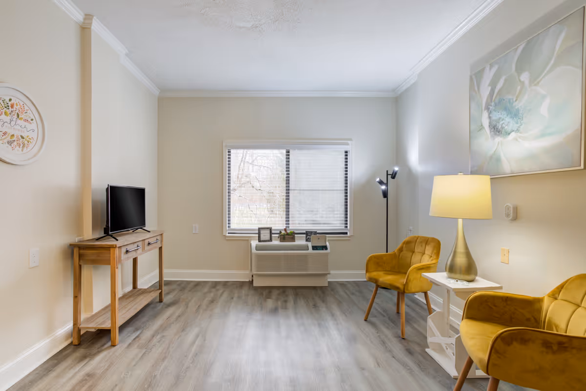 Bright living room with two mustard-yellow armchairs, a lamp and side table, a TV on a wooden console, and a window with an air conditioning unit.