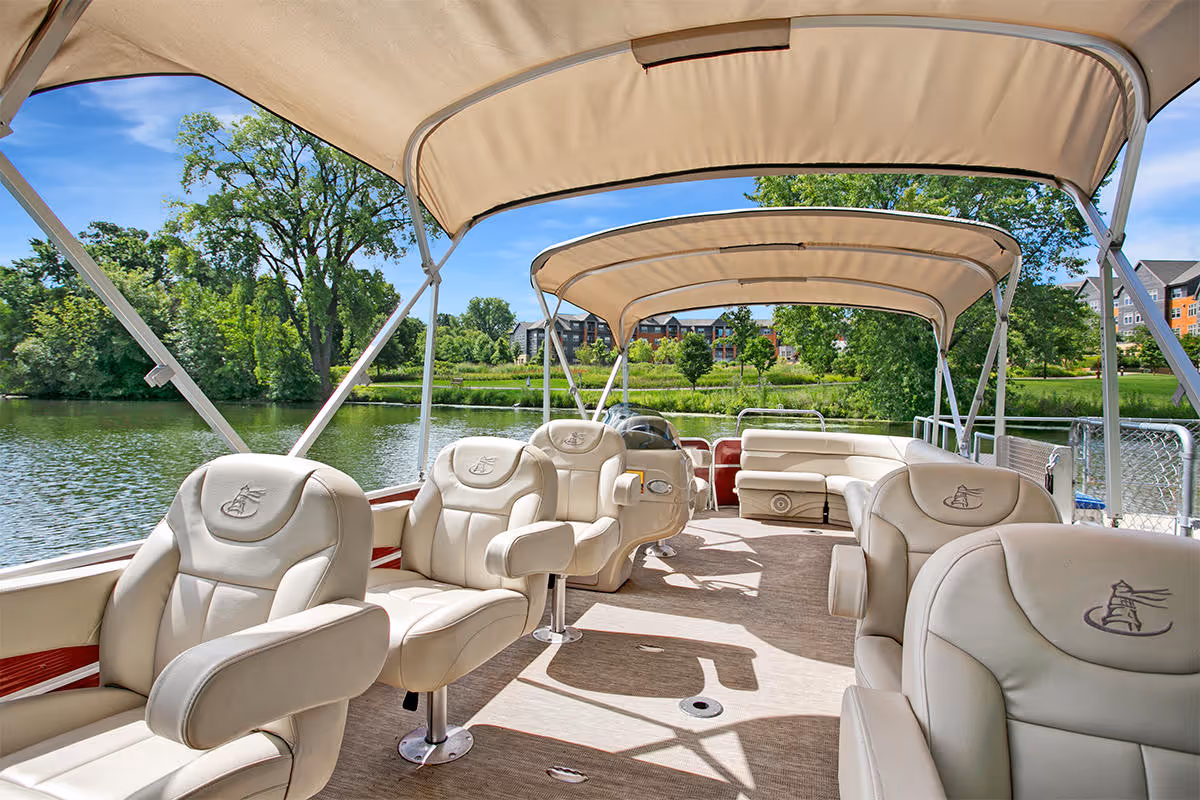Covered pontoon boat deck with cream leather captain chairs under tan canopies on a lake with trees and buildings in the background.