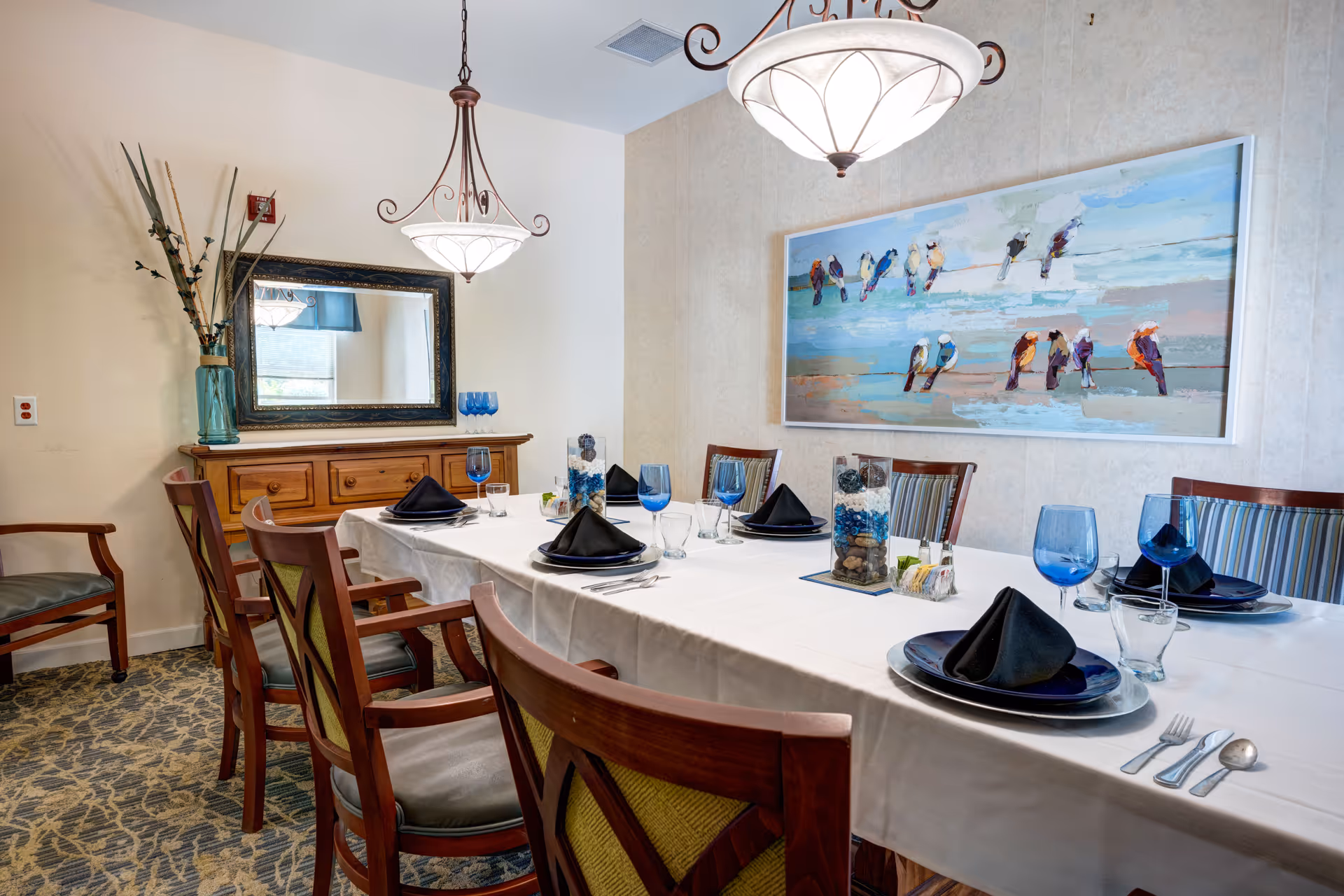 Elegantly set dining room table with wooden chairs, blue glassware, black napkins, and a coastal bird painting on the wall.