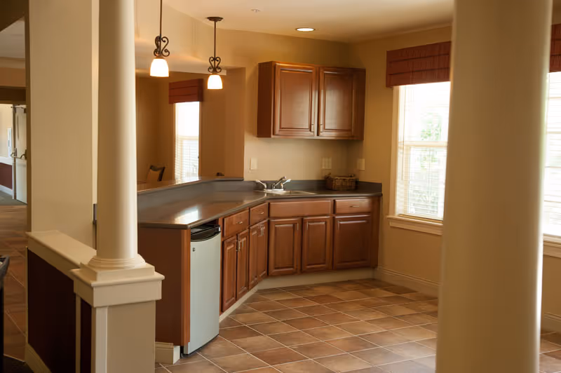 Interior view of a kitchenette area with wooden cabinets, a small refrigerator, a sink, and tiled flooring. Two pendant lights hang from the ceiling, and there are windows with blinds letting in natural light.