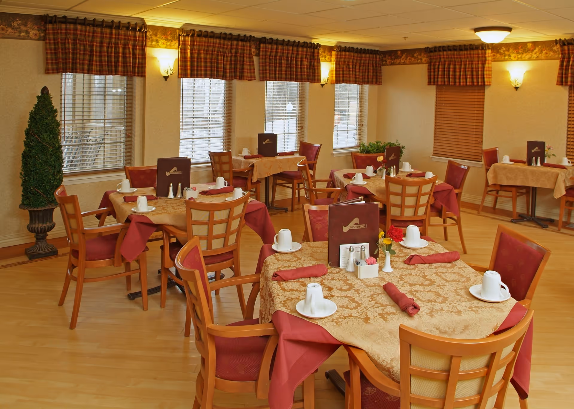 A dining room in a senior living community with several tables covered in beige and burgundy tablecloths. Each table is set with white cups and saucers, red cloth napkins, salt and pepper shakers, and small flower arrangements. The room has wooden chairs with red cushions, windows with blinds and plaid valances, and warm wall sconces providing lighting.