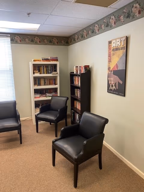 A small room with three black leather chairs arranged around the space. There are two bookshelves filled with books against the walls, one white and one black. A window with blinds is on the left side, and a vintage-style poster about art hangs on the right wall. The room has beige carpet and floral wallpaper border near the ceiling.