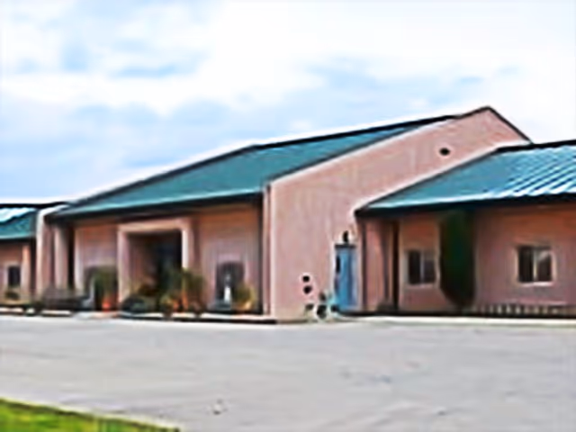 Exterior view of a single-story building with a green metal roof and light-colored walls, featuring multiple entrances and small windows, set against a partly cloudy sky.