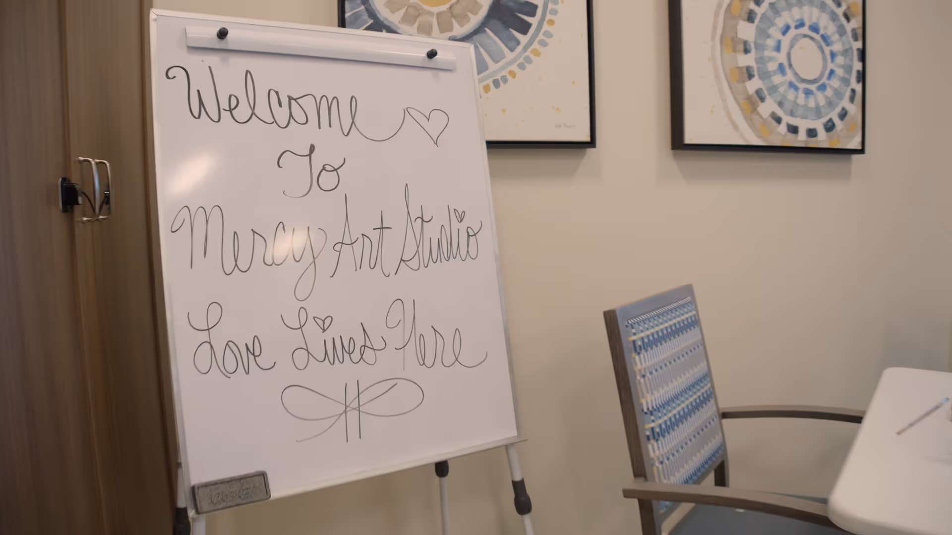 A whiteboard with handwritten text that says 'Welcome to Mercy Art Studio Love Lives Here' with hearts and decorative lines. The whiteboard is placed next to a wooden cabinet and a chair with a blue patterned backrest. Two framed abstract artworks hang on the beige wall behind.