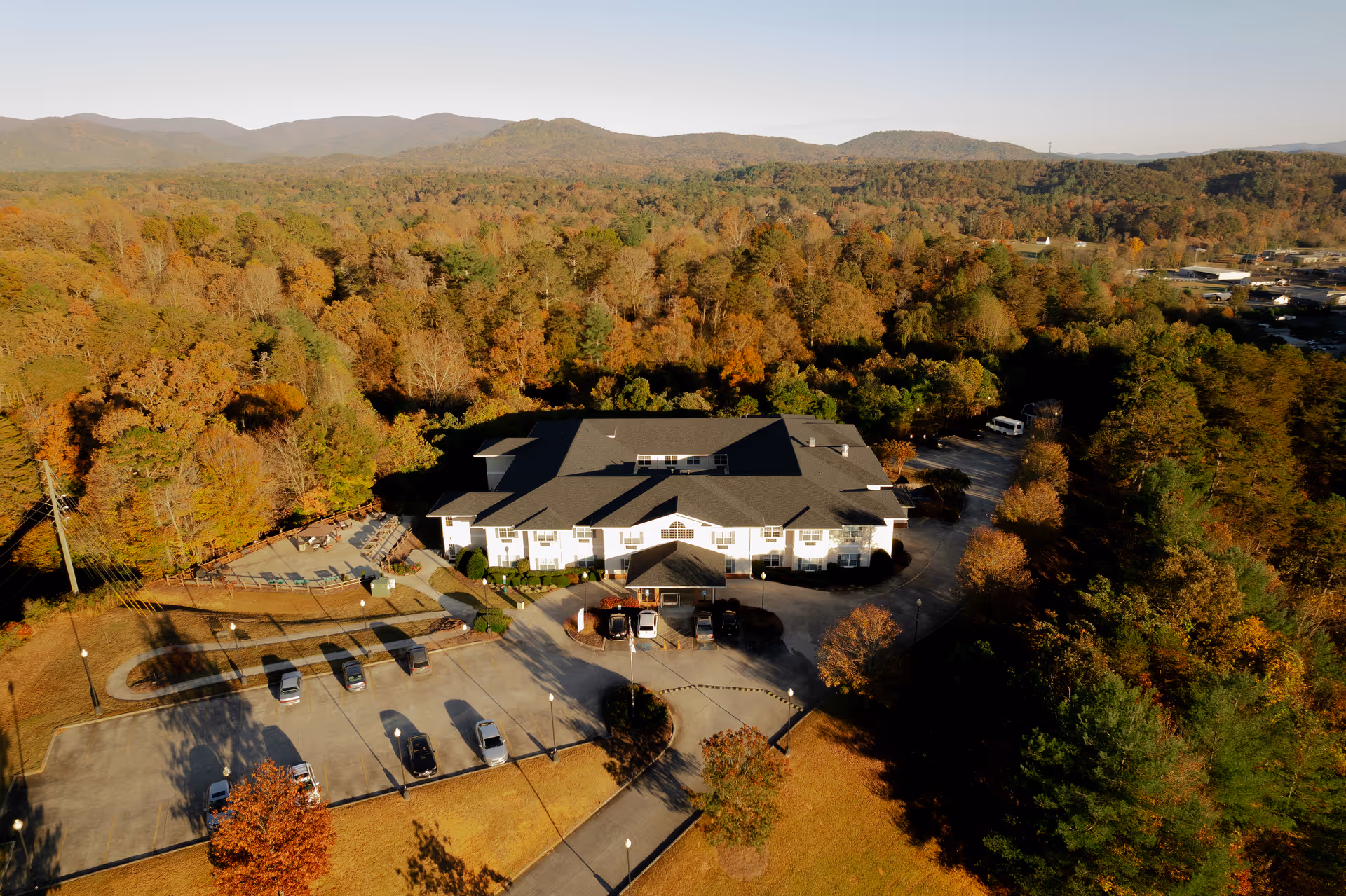 Aerial view of a two-story senior living building surrounded by autumn trees, a parking lot, and distant mountains.