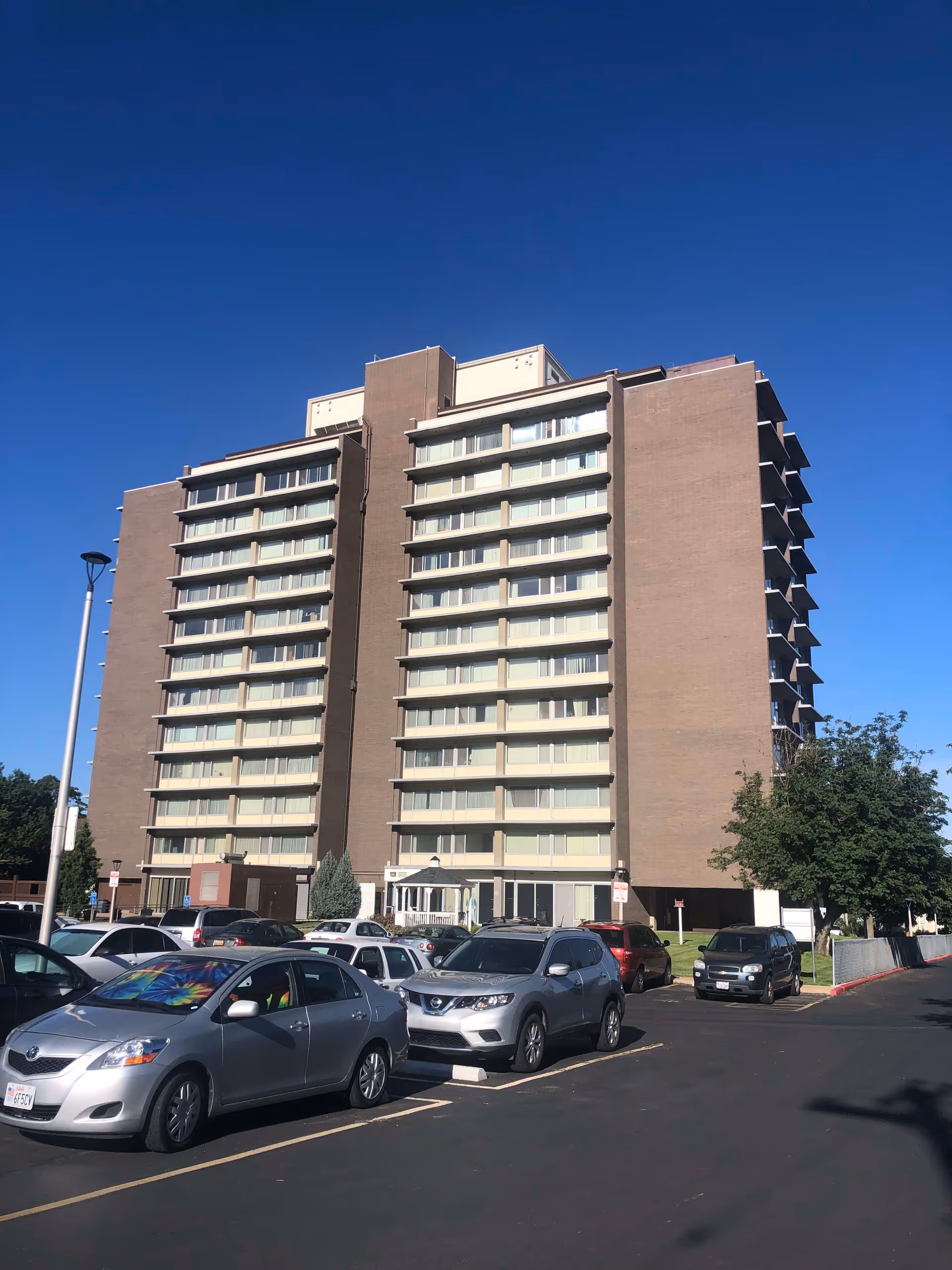 Tall multi-story brick residential building with balconies and a parking lot of cars under a clear blue sky.
