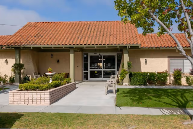 Front entrance of Camarillo Healthcare Center with a tiled roof, beige walls, a covered porch area with benches, a small garden bed with plants, and a tree on the right side under a clear blue sky.