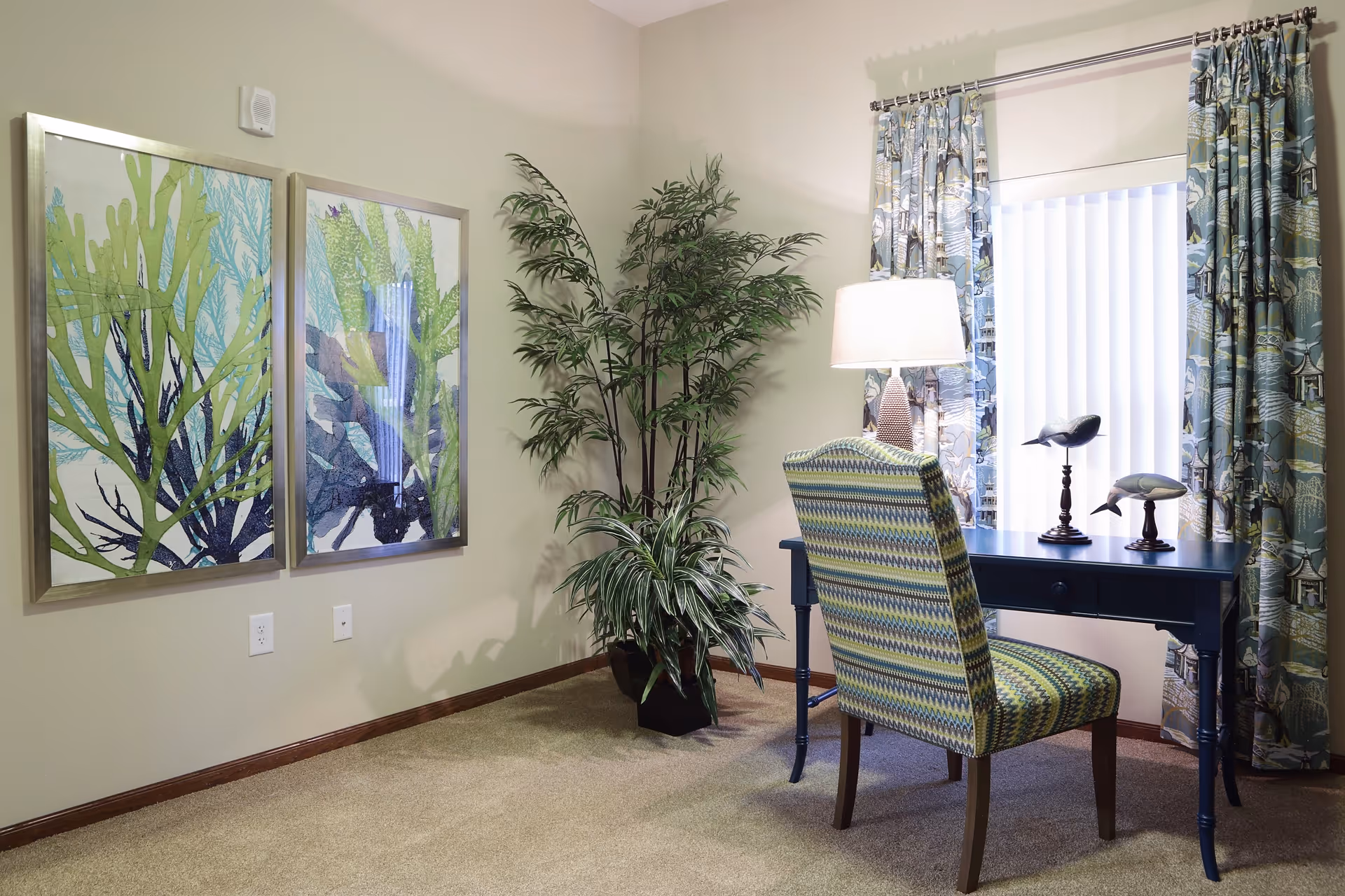 A cozy corner of a room featuring a patterned upholstered chair in front of a dark blue desk with two decorative bird figurines and a lit table lamp. The window behind the desk is covered with vertical blinds and patterned curtains. On the left wall, there are two framed artworks depicting green and blue seaweed or coral. A tall green potted plant is placed in the corner between the desk and the artwork.