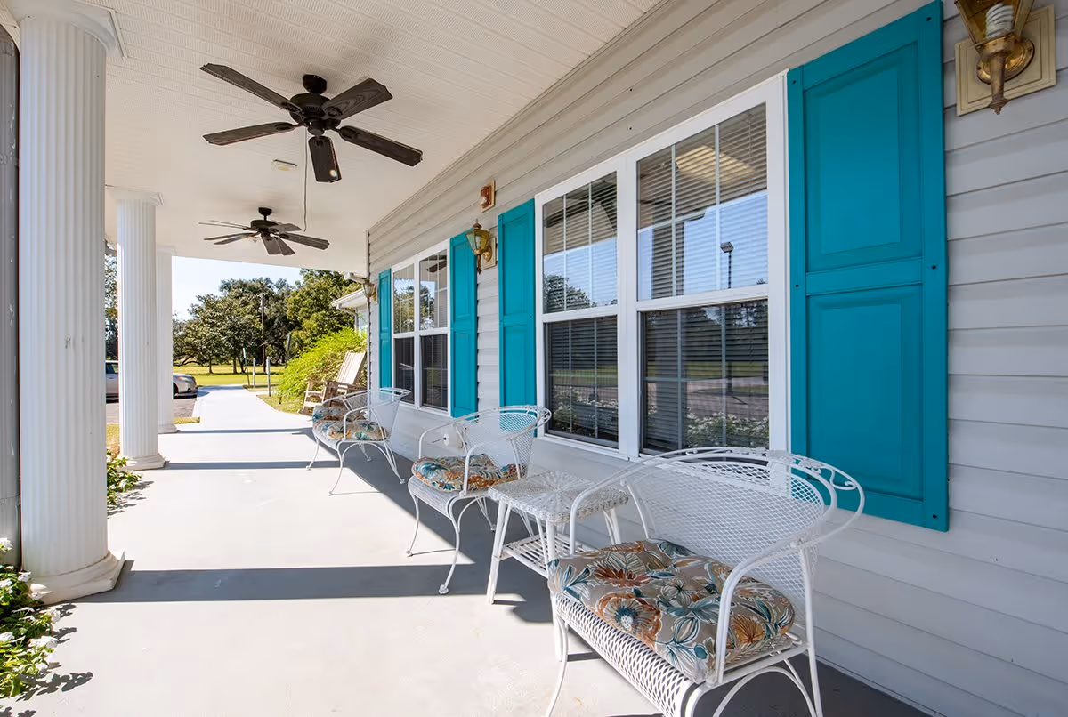 A covered outdoor porch area with white wicker chairs featuring floral cushions, a small white wicker table, and two ceiling fans. The porch has large white columns and blue window shutters on the building's exterior wall. Trees and a parking area are visible in the background.