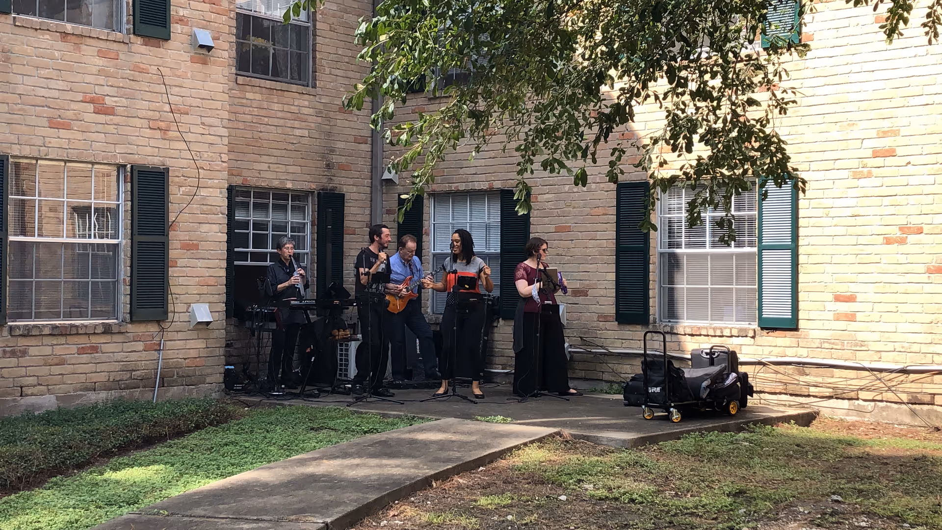 A small band of five musicians performing in a brick courtyard beside a residential building.