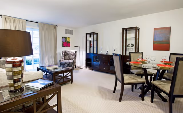 Bright furnished living and dining area with sofas and armchair, glass dining table set for four, dark wood cabinets and a large curtained window.