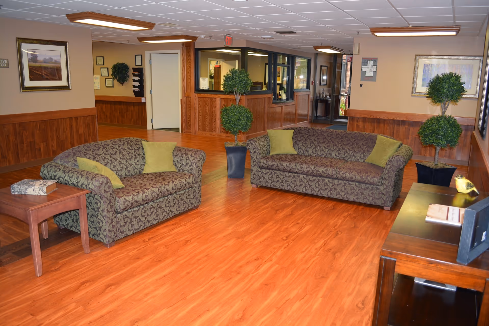 A nursing facility common area with two patterned sofas each adorned with green throw pillows, a wooden side table with a decorative box, two potted topiary plants, wood-paneled walls, framed artwork, and a wooden floor. The area is well-lit with ceiling lights and has a reception window and an exit door in the background.