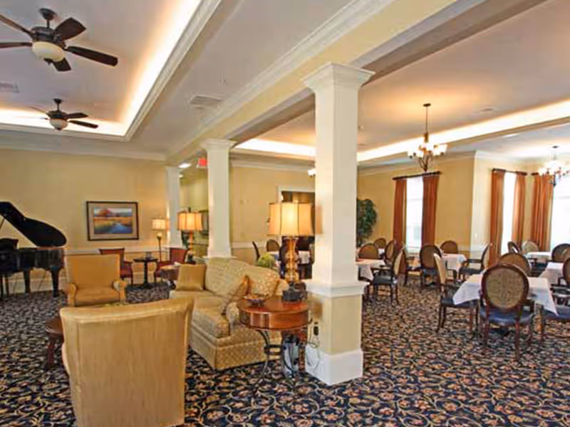 A spacious and elegant common area in a senior living facility featuring comfortable seating with armchairs and a sofa, a grand piano, several tables with chairs covered in white tablecloths, decorative lamps, ceiling fans, and large windows with curtains allowing natural light to fill the room.