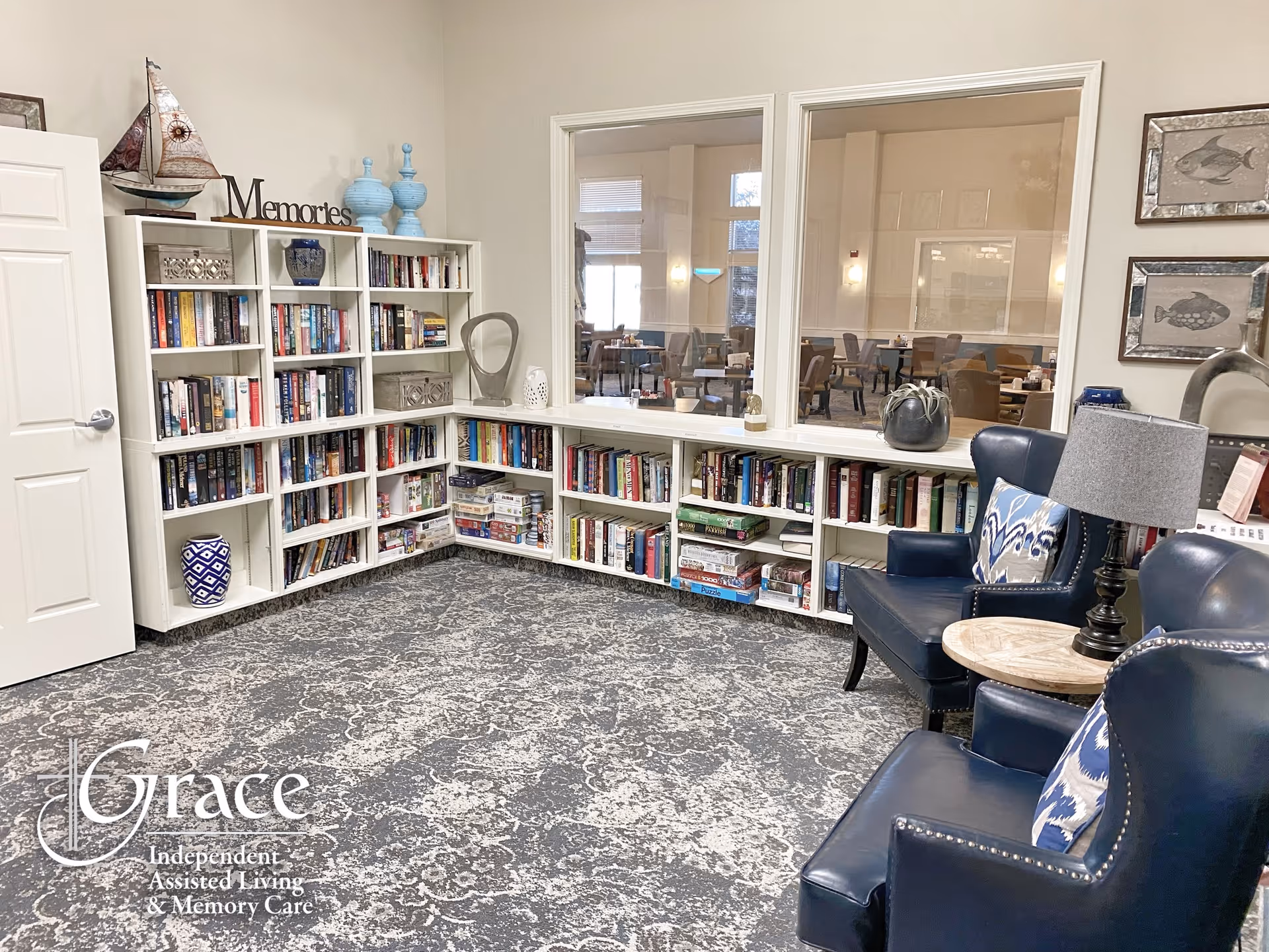 A cozy reading nook in an assisted living facility featuring white bookshelves filled with books and board games, two blue leather armchairs with patterned pillows, a small round wooden side table with a lamp, decorative items including a model sailboat and vases, and a large window looking into a dining area.