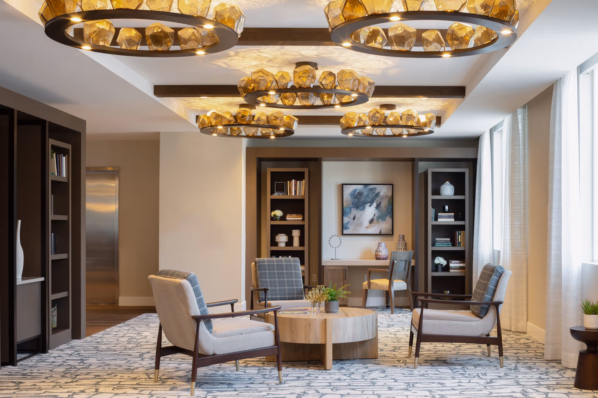 A cozy senior living common area with four cushioned armchairs arranged around a round wooden coffee table. The room features patterned carpet, built-in bookshelves with books and decorative items, a desk with a chair, abstract wall art, and large windows with sheer curtains allowing natural light. The ceiling has wooden beams and multiple modern circular chandeliers with amber-colored glass accents.