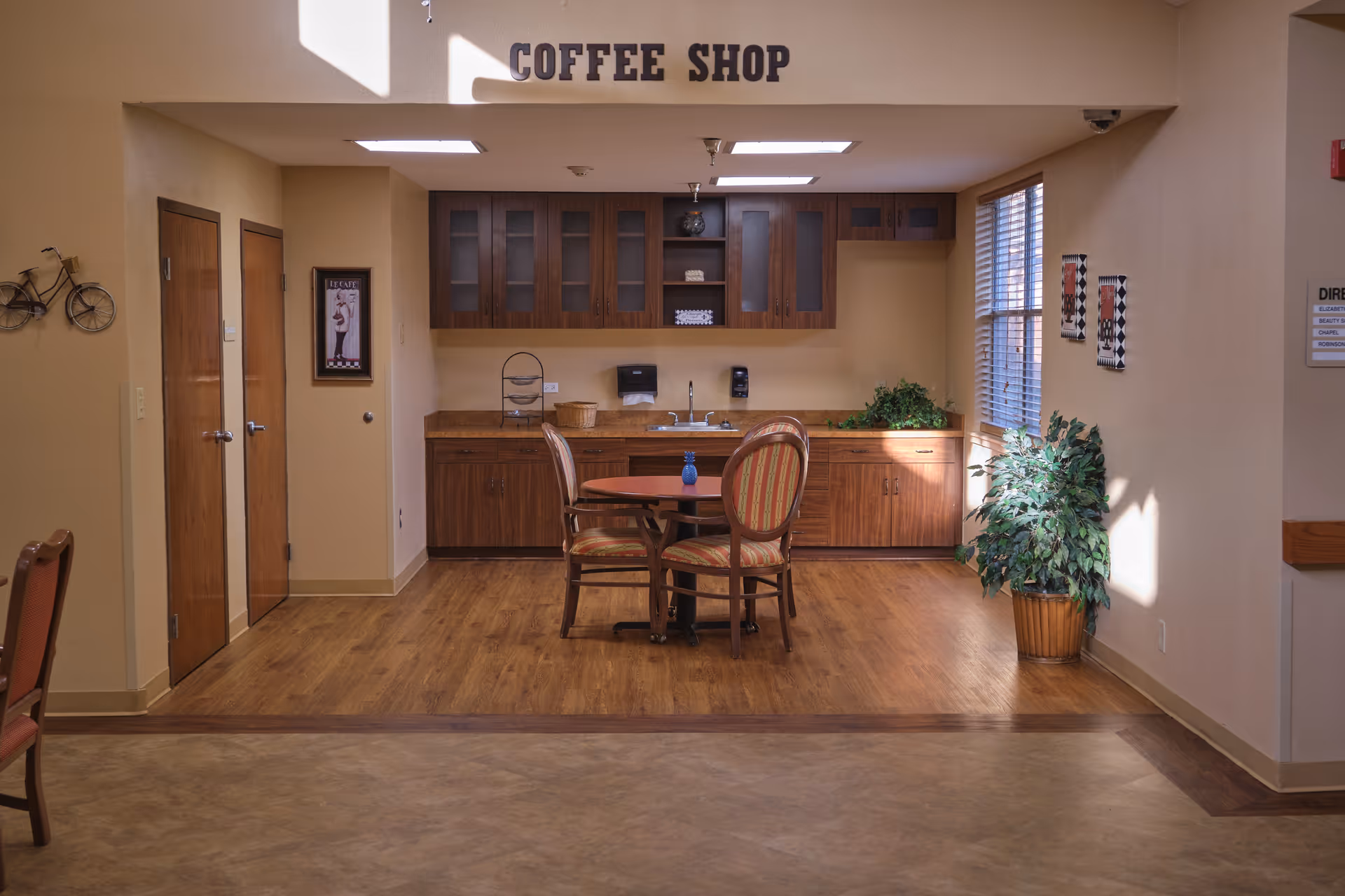 Interior view of a coffee shop area in a senior living facility with wooden cabinets, a countertop with a sink, and a small round table with three chairs. There is a large potted plant near a window with blinds, wall decorations, and a sign above the area that reads 'COFFEE SHOP'.