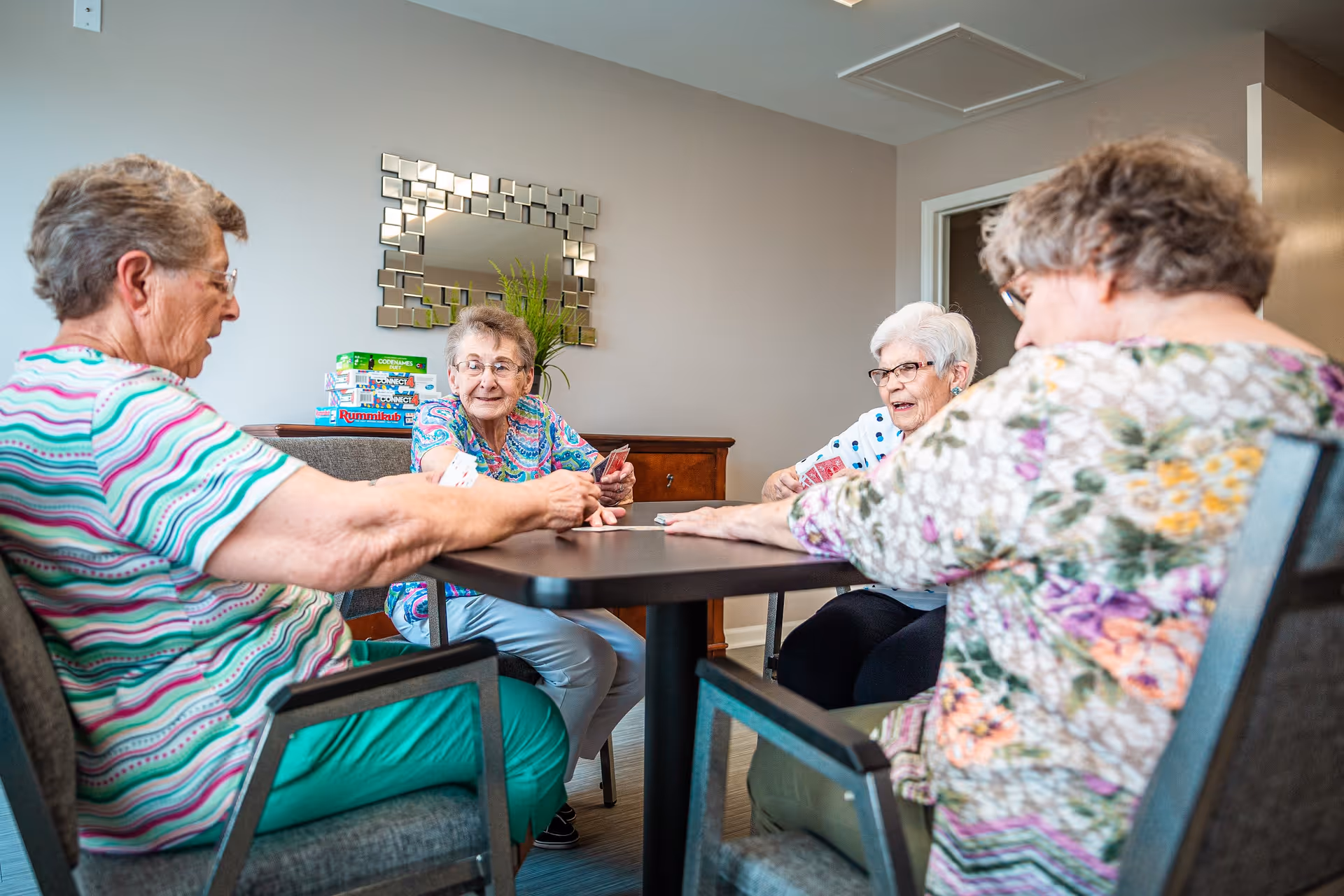 Four elderly women sitting around a square table playing a card game in a well-lit room with a decorative mirror and a plant on a wooden sideboard in the background.