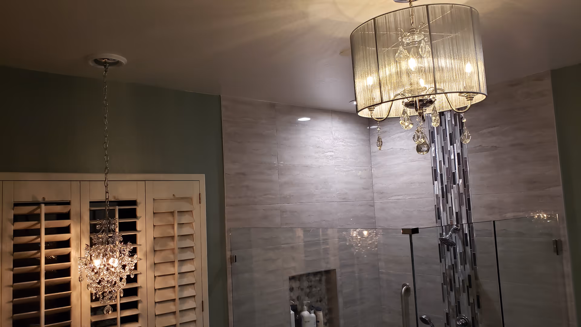 Modern bathroom shower area with glass enclosure, tiled walls featuring a vertical mosaic strip, and two elegant crystal chandeliers hanging from the ceiling. There are white wooden shutters on the left side.