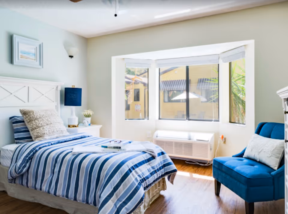 Bright bedroom with a single bed in blue-striped linens, a nightstand and lamp, bay windows, and a blue armchair.