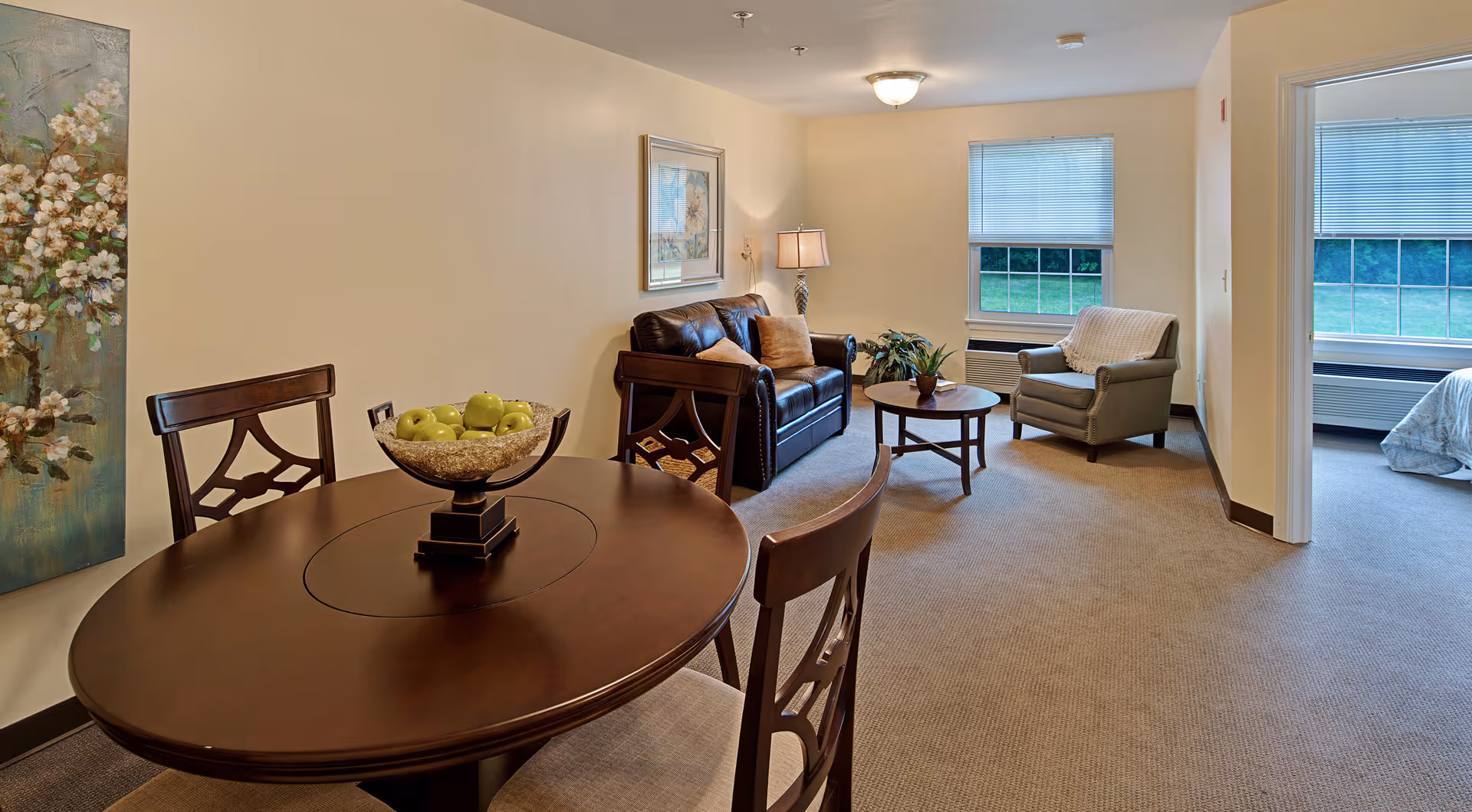A cozy senior living area featuring a round wooden dining table with a bowl of green apples, two wooden chairs, a brown leather sofa with cushions, a small round coffee table with a plant, and a comfortable armchair. The room has beige walls, carpeted floor, two windows with blinds, and a glimpse of a bedroom with a bed visible through an open doorway.