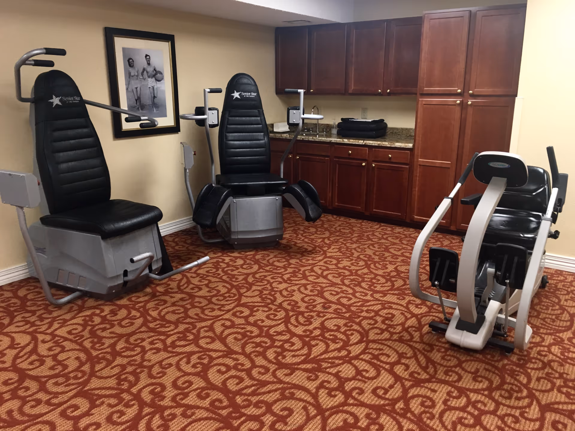 Small exercise room with three seated/resistance machines, wood cabinets and a patterned carpeted floor.