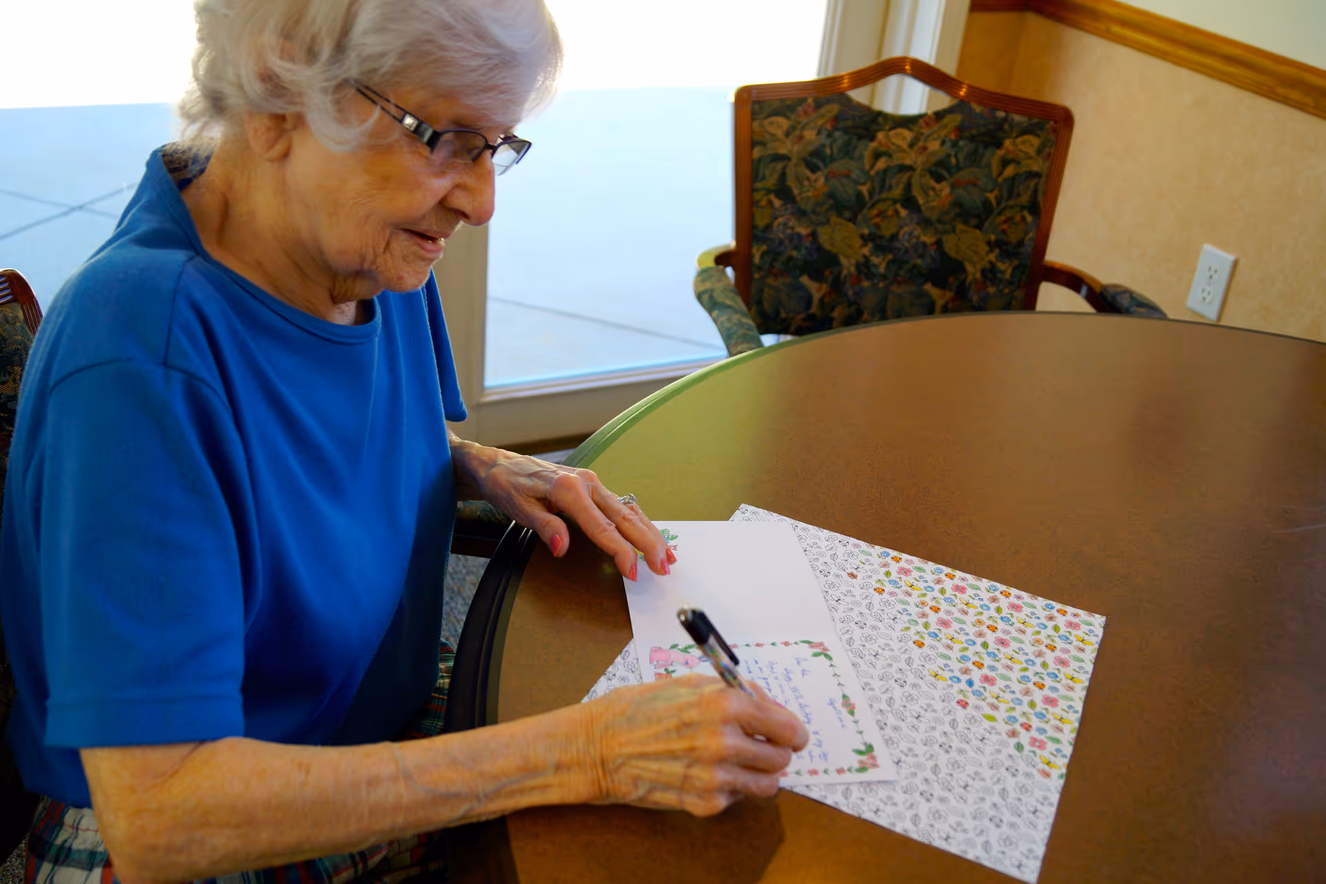 An elderly woman with white hair and glasses is sitting at a round table in a room, writing on a decorative card with a pen. The table has a floral placemat, and there is a floral upholstered chair nearby. A window or glass door is visible in the background.