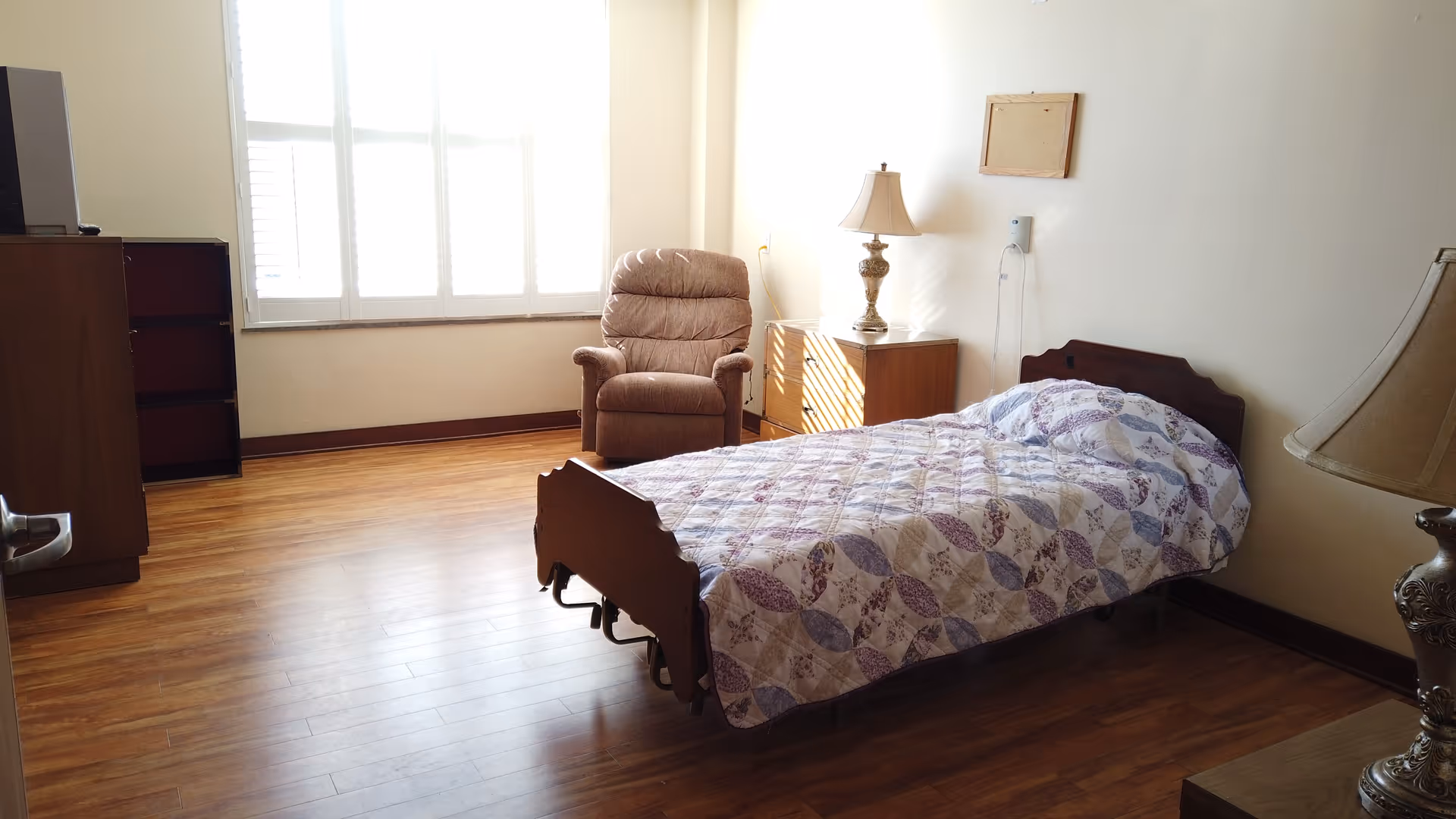 A simple bedroom with a single bed covered with a patterned quilt, a cushioned armchair near a window with white blinds, two wooden nightstands each with a decorative lamp, and wooden flooring.