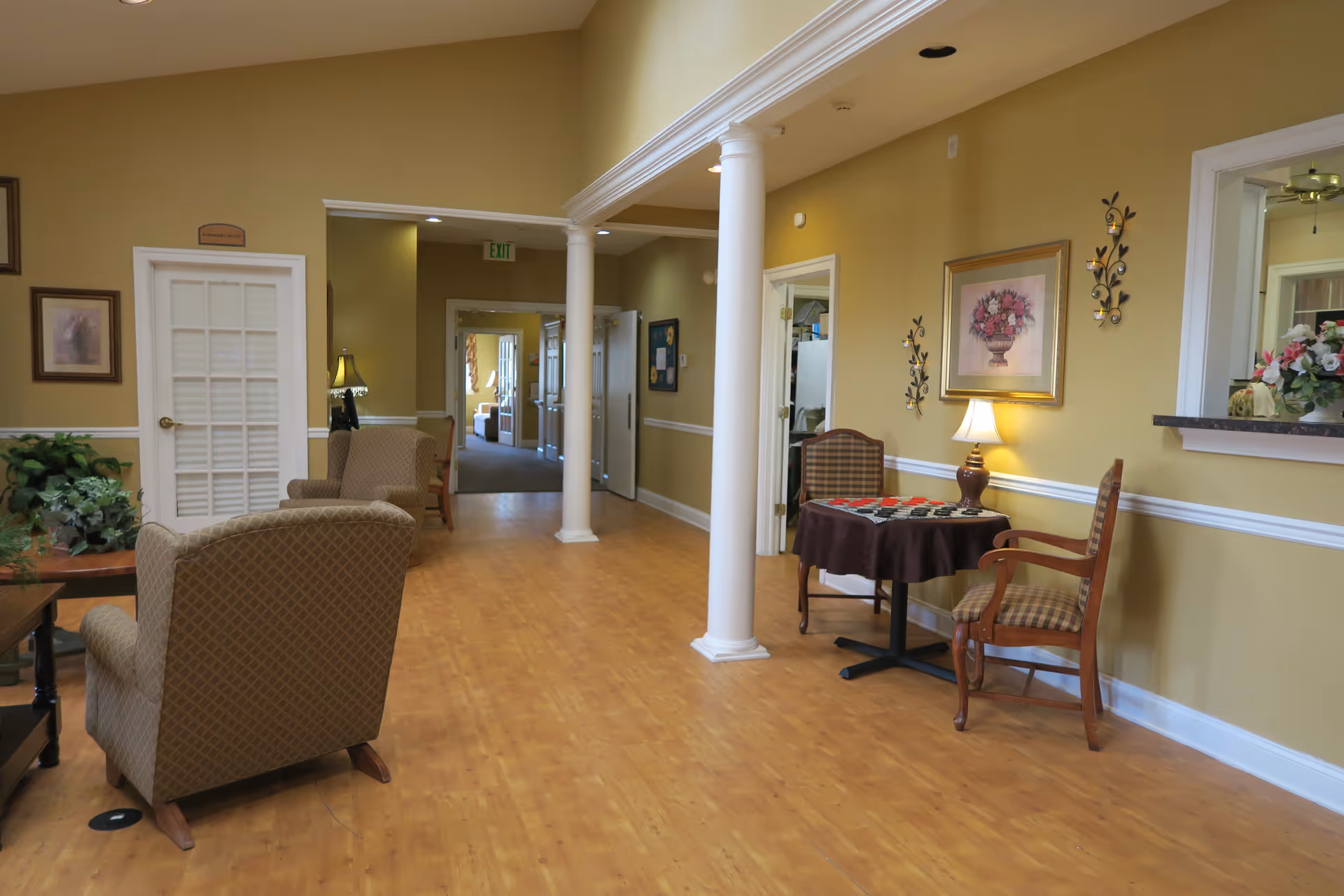 Interior view of a senior living facility hallway with beige walls and wooden flooring. The space features comfortable armchairs, a small table with two chairs and a lamp, decorative wall art, and white columns. There is a doorway with a glass-paneled door and an exit sign visible in the background.
