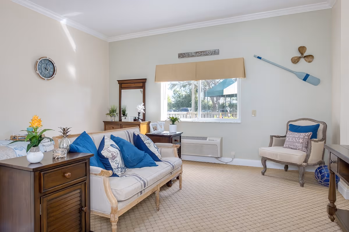 A cozy living room with a beige sofa adorned with blue and patterned pillows, a wooden side table with a small plant, a wooden dresser with a mirror, and a beige armchair with decorative pillows. The room has a large window with a beige valance, a wall clock, a decorative wooden oar, and a small propeller mounted on the wall. The carpet is light beige with a subtle pattern.