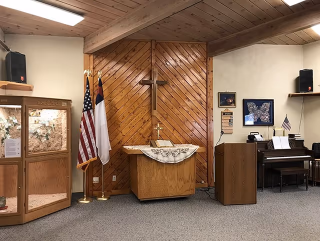 Interior room with a wooden podium draped with a white cloth, a wooden cross mounted on a wood-paneled wall behind it, and two flags (American and Christian) standing to the left. To the right, there is a wooden lectern and a piano with sheet music and a small American flag on top. The room has a carpeted floor and beige walls.