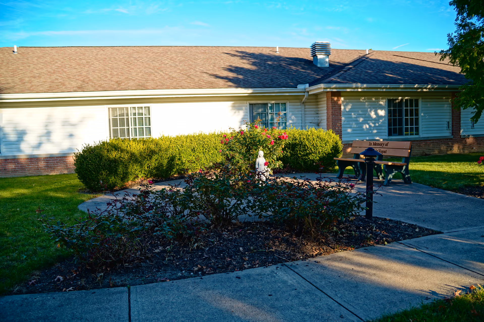 Outdoor garden area with a concrete pathway, bushes, and a small statue in the center surrounded by flowers. There is a bench with a memorial plaque and a building with white siding and brick accents in the background under a clear blue sky.