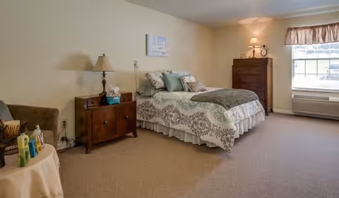A spacious bedroom with a bed covered in patterned bedding and multiple pillows. There is a wooden nightstand with a lamp and tissues next to the bed, a wooden dresser with a lamp and clock, a window with a valance, and a beige carpeted floor. A small table with decorative items and a cushioned chair are also visible.