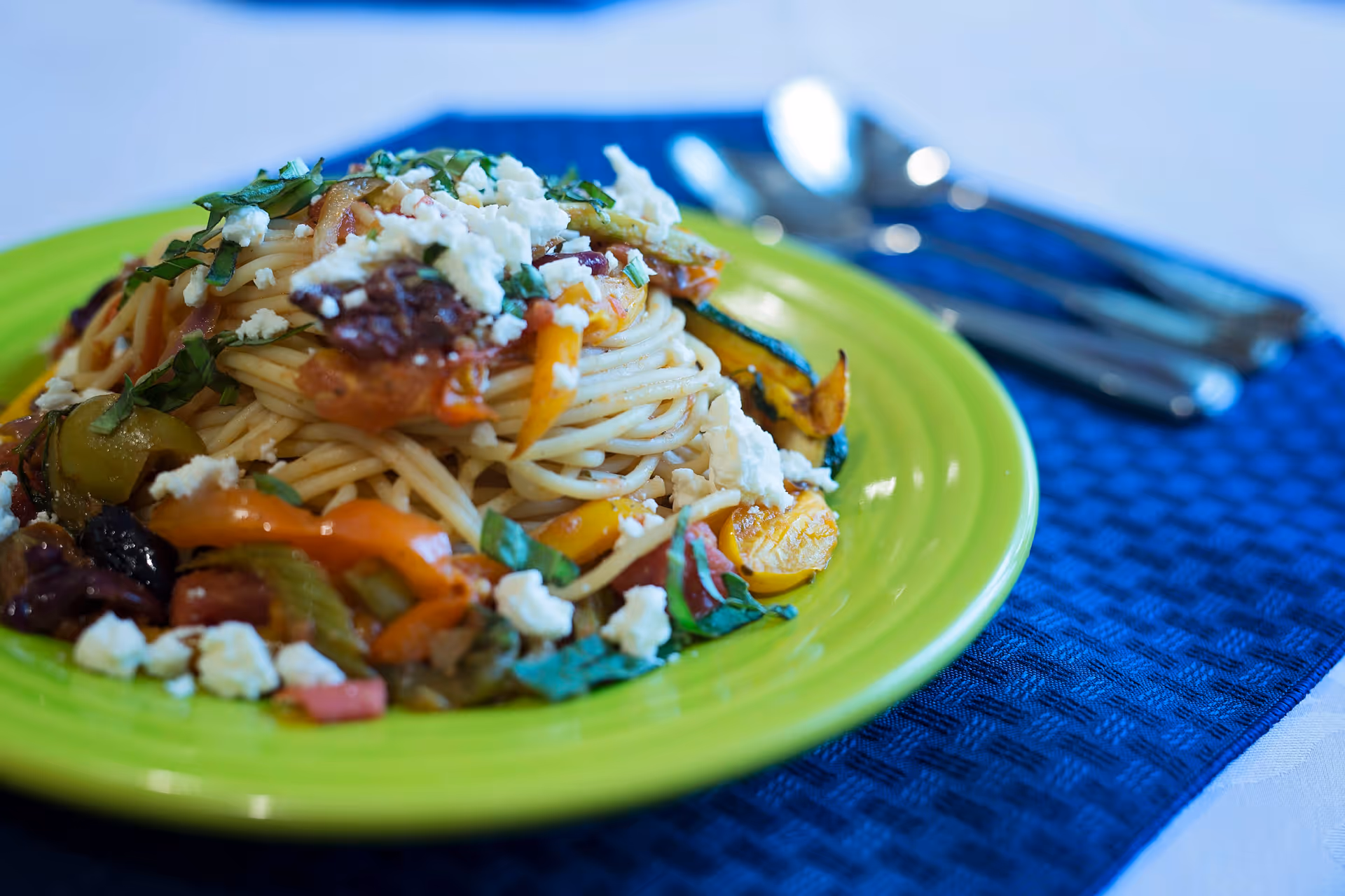 A plate of spaghetti pasta topped with crumbled cheese, chopped herbs, and mixed sautéed vegetables on a green plate, placed on a blue textured placemat with silverware in the background.
