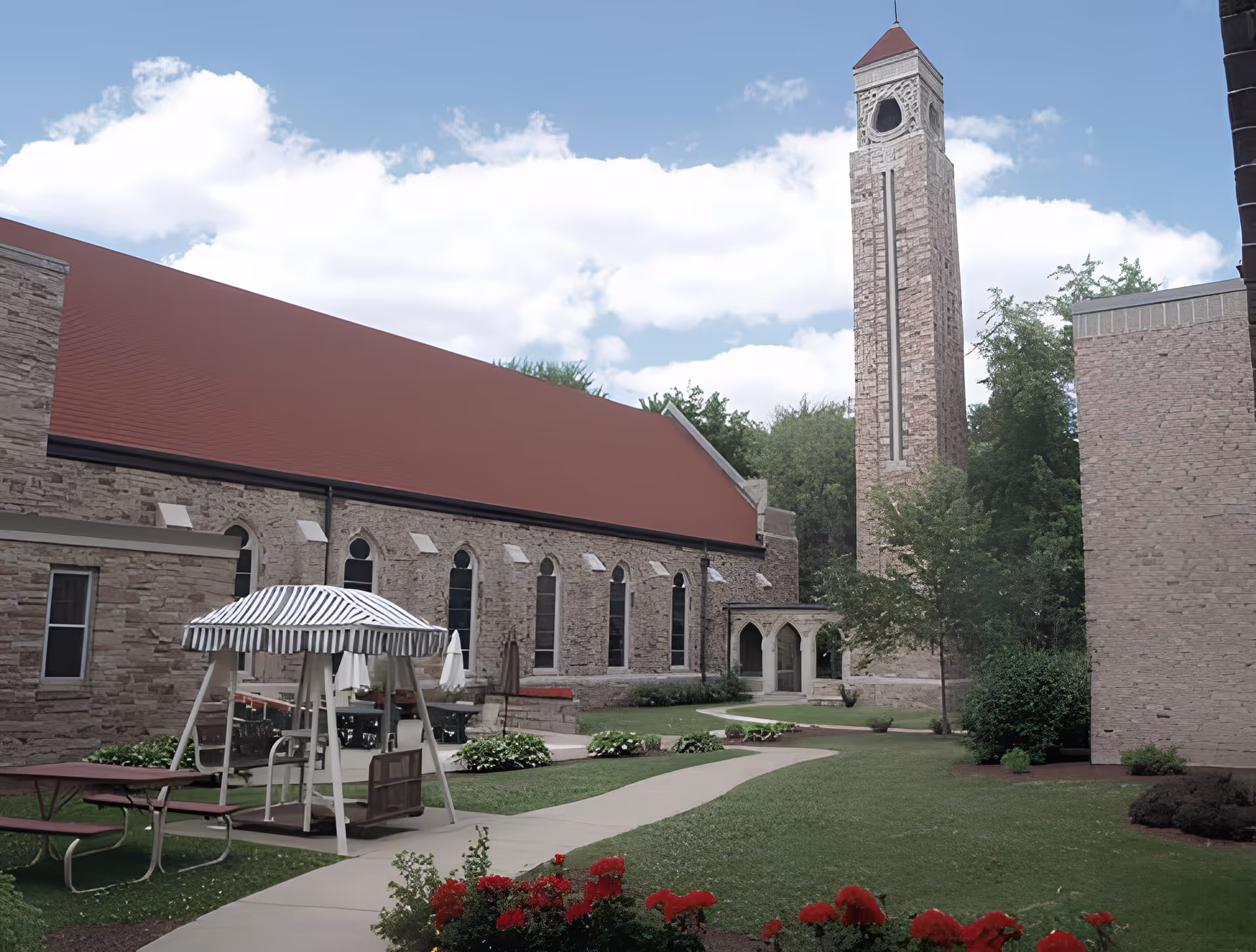 Outdoor courtyard area with a stone building featuring arched windows and a tall clock tower. There is a paved walkway, green grass, red flowers, a striped canopy swing, and picnic tables. Trees and bushes surround the area under a partly cloudy sky.