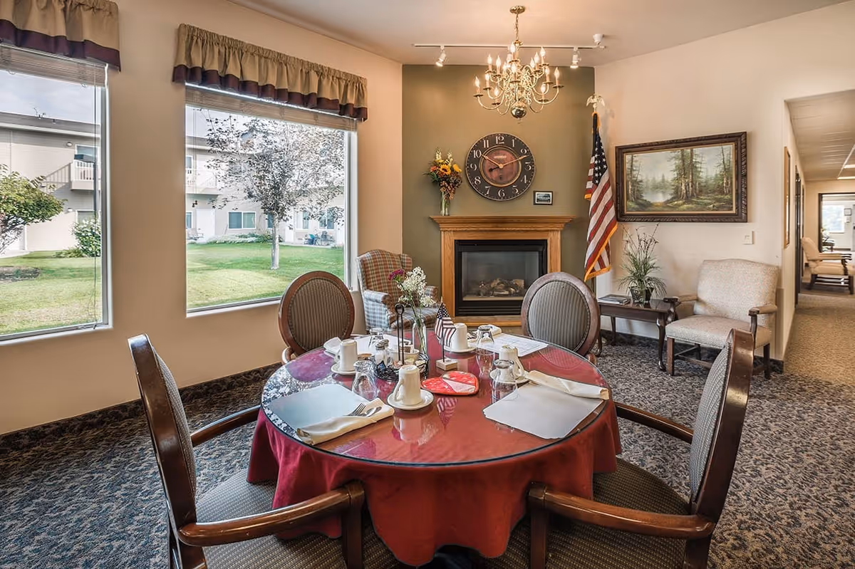 Round dining table set for a meal in a cozy common room with a fireplace, chandelier, and large windows overlooking a courtyard.