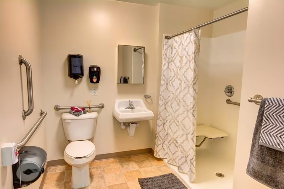 Accessible bathroom with a toilet, wall-mounted sink, grab bars, and a shower with curtain and fold-down seat.