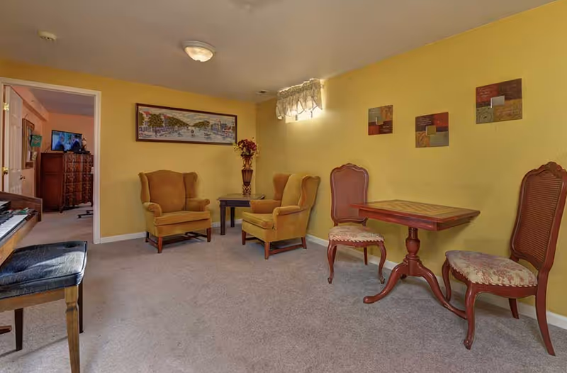 A cozy sitting room with yellow walls featuring two mustard armchairs around a small side table, a small wooden table with two chairs, and a piano bench near a doorway.
