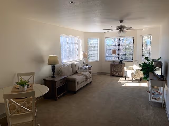 A bright living room with beige carpet and white walls featuring a small round dining table with two chairs, a beige loveseat with a 'HOME' pillow, two side tables with lamps, a patterned armchair near large windows with blinds, a small TV on a white stand, and a green plant beside the TV stand.