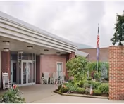 Covered front entrance of a senior living facility with rocking chairs, landscaped beds, and an American flag.