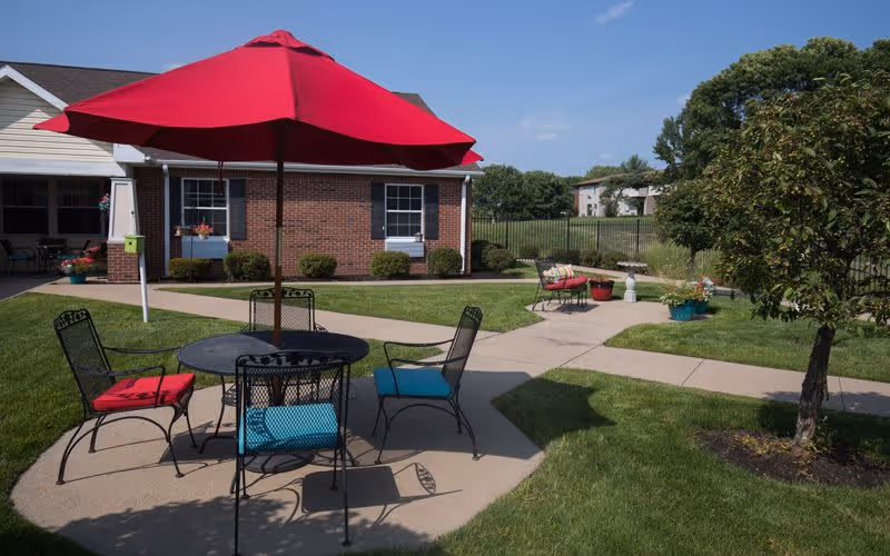 Outdoor courtyard with a round patio table, chairs with colorful cushions and a red umbrella in front of a brick building.