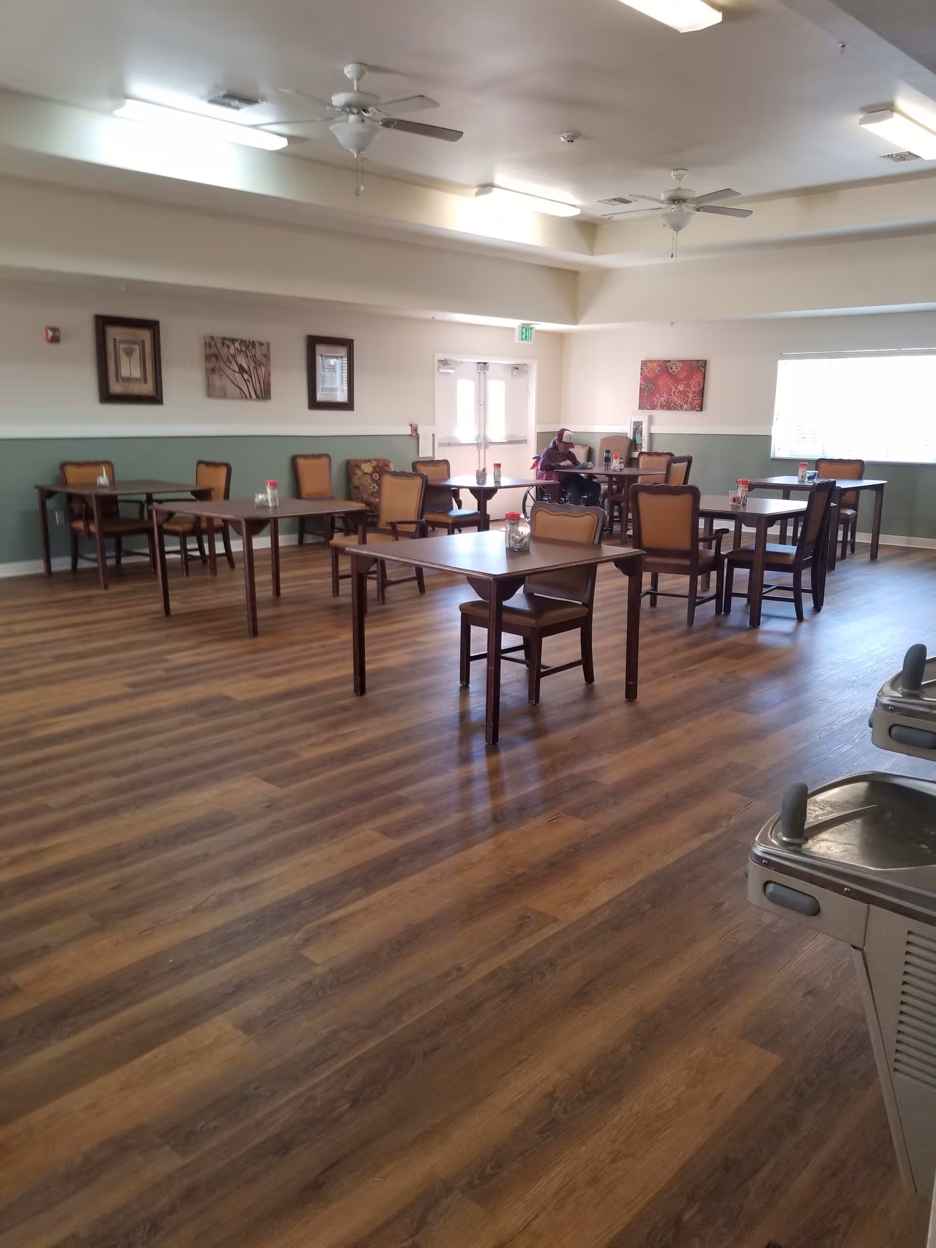 A spacious dining room with several wooden tables and chairs arranged neatly. The room has wood-patterned flooring, light-colored walls with green wainscoting, and ceiling fans. There are framed pictures on the walls and a large window letting in natural light. A person in a wheelchair is seated at one of the tables near the back of the room.